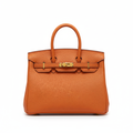 An orange pebbled leather handbag with gold-tone hardware, featuring dual top handles and a front strap closure, photographed against a solid white background.